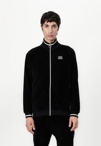 Giacca nera in corduroy con zip e colletto alto, dotata di una zip bianca, polsini a righe e un piccolo logo sul lato sinistro del petto.