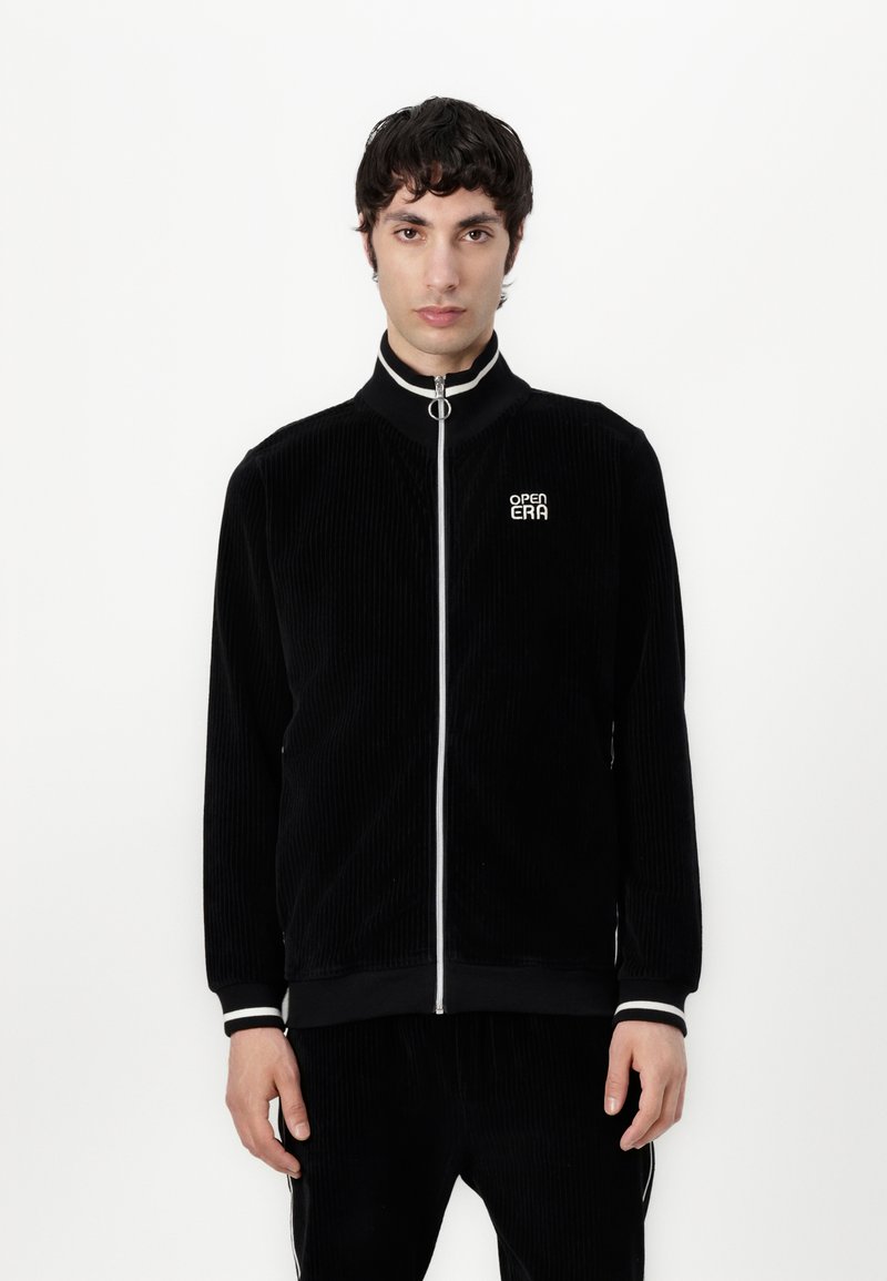 Giacca nera in corduroy con zip e colletto alto, dotata di una zip bianca, polsini a righe e un piccolo logo sul lato sinistro del petto.