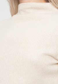 Μπεζ, ριγωτό πλεκτό πουλόβερ με υψηλό λαιμό. Το ύφασμα φαίνεται μαλακό και υφασμάτινο, με φωτεινό, ουδέτερο χρώμα.