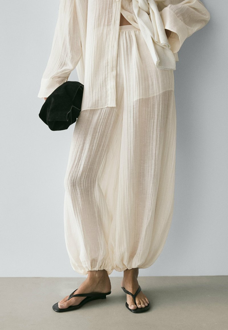 Person iført gennemsigtige, løstsiddende cremefarvede bukser og matchende top, der holder en sort hat, med sorte flip-flops på bare fødder.