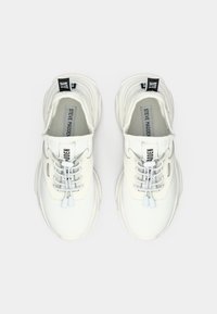 Balti Steve Madden sportbačiai su tekstūruota medžiaga, bungee tipo raišteliais ir juodomis kilpomis ant paprasto balto fono.