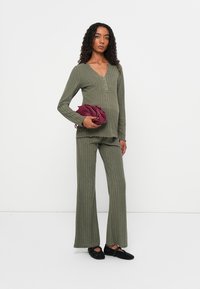 Olivgrünes, geripptes Stricktop mit V-Ausschnitt und Knöpfen, kombiniert mit passenden, ausgestellten Hosen. Dazu eine kastanienbraune Clutch und schwarze Schuhe.