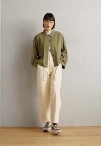 Oliven grønn kort jakke med krage og frontknapper, kombinert med lyse beige bukser og grå sneakers. Glatt tekstur, uformell design.