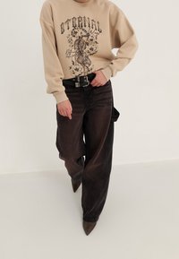 Sweat-shirt beige avec un motif graphique de tigre et de fleurs. Jean noir à jambes larges avec une ceinture noire et des talons pointus marron.