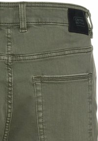 Olive green denim pants feature a black leather brand patch, back pocket stitching, and a standard waistband with belt loops.