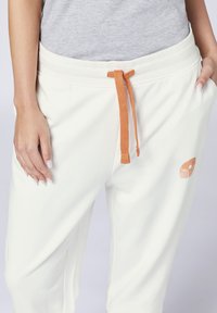 Weiße Baumwoll-Jogginghosen mit einem elastischen Bund, orangefarbener Kordel und einem kleinen grafischen Detail am Oberschenkel. Weicher Stoff und lockere Passform.