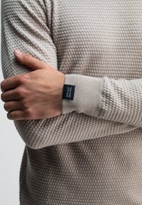 Man in een beige gebreide trui met textuur en een zwart "BUTCHER OF BLUE" label op de manchette, die zijn tegenovergestelde arm tegen een effen achtergrond houdt.