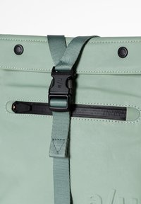 Zaino di tessuto verde con una superficie liscia, dotato di una fibbia nera, cinturino regolabile e apertura con zip nella parte superiore.
