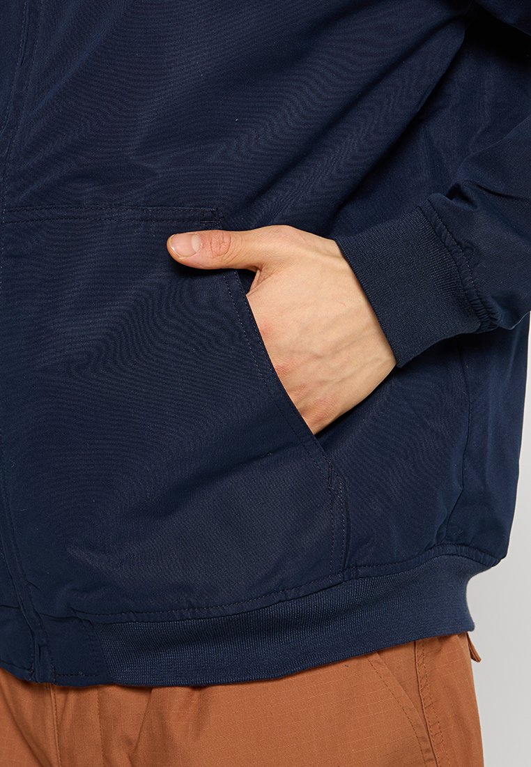 Granatowa kurtka o gładkiej teksturze, elastycznych mankietach i widocznej kieszeni bocznej. Prosty design z luźnym krojem.