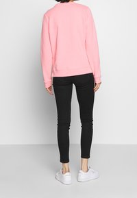 Person in einem hellrosa langärmeligen Sweatshirt, schwarzen Skinny Jeans und weißen Reebok-Sneakers, der vor einem schlichten Hintergrund steht.