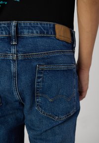 Kék farmer nadrág klasszikus szabással, barna bőr folttal, amelyen az "American Eagle" márkajelet találhatjuk, valamint jellegzetes varrással a hátsó zsebeken.