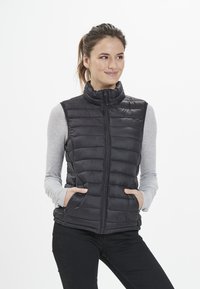 Nuori nainen, jolla on ruskeat hiukset poninhännällä, pukeutuneena mustaan tikattuun hihattomaan liiviin vaalean harmaan pitkähihaisen paidan päällä, kädet taskuissa.