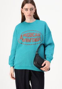 Sweatshirt oversized turquesa com "AMERICAN VINTAGE" impresso em castanho. Combinado com uma mala de couro preta e calças riscas de giz.
