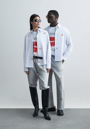 Zwei Models tragen aufeinander abgestimmte Outfits: ein weißes Hemd mit Lippenprint, grafische T-Shirts, graue Jogginghosen und schwarze Stiefel mit Absatz.