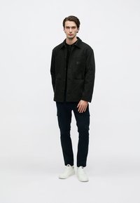 Μαύρο υφασμάτινο μπουφάν με κουμπιά στο μπροστινό μέρος, δύο τσέπες μπροστά και υφασμένο γιακά, συνδυασμένο με σκούρα cargo παντελόνια και λευκά αθλητικά παπούτσια.