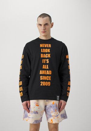 Long-sleeve black T-shirt with orange text reading "NEVER LOOK BACK IT’S ALL AHEAD SINCE 2009." Paired with pastel, splatter-pattern shorts.