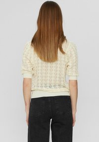 Pletený krémový svetr s texturovaným vzorem listů. Krátké rukávy a žebrovaný spodní lem; v kombinaci s černými džíny s vysokým pasem.