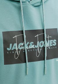Türkiisikainega kapuutsiga dressipluus, millel on triibuline must ristkülikukujuline logomärk, millel on kiri "Jack & Jones Denim" ning sobivad lintnöörid täpilise mustriga.