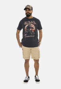 Camiseta gráfica negra con diseño de motocicleta y lobo, pantalones cortos de carga caqui y zapatillas negras con suelas blancas. La gorra combina con los colores de la camiseta.