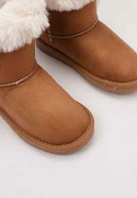 Botas de tobillo de ante marrón con un suave puño de piel de piel, forma de puntera redonda y suela de goma texturizada. Detalle de costura visible a lo largo del borde.