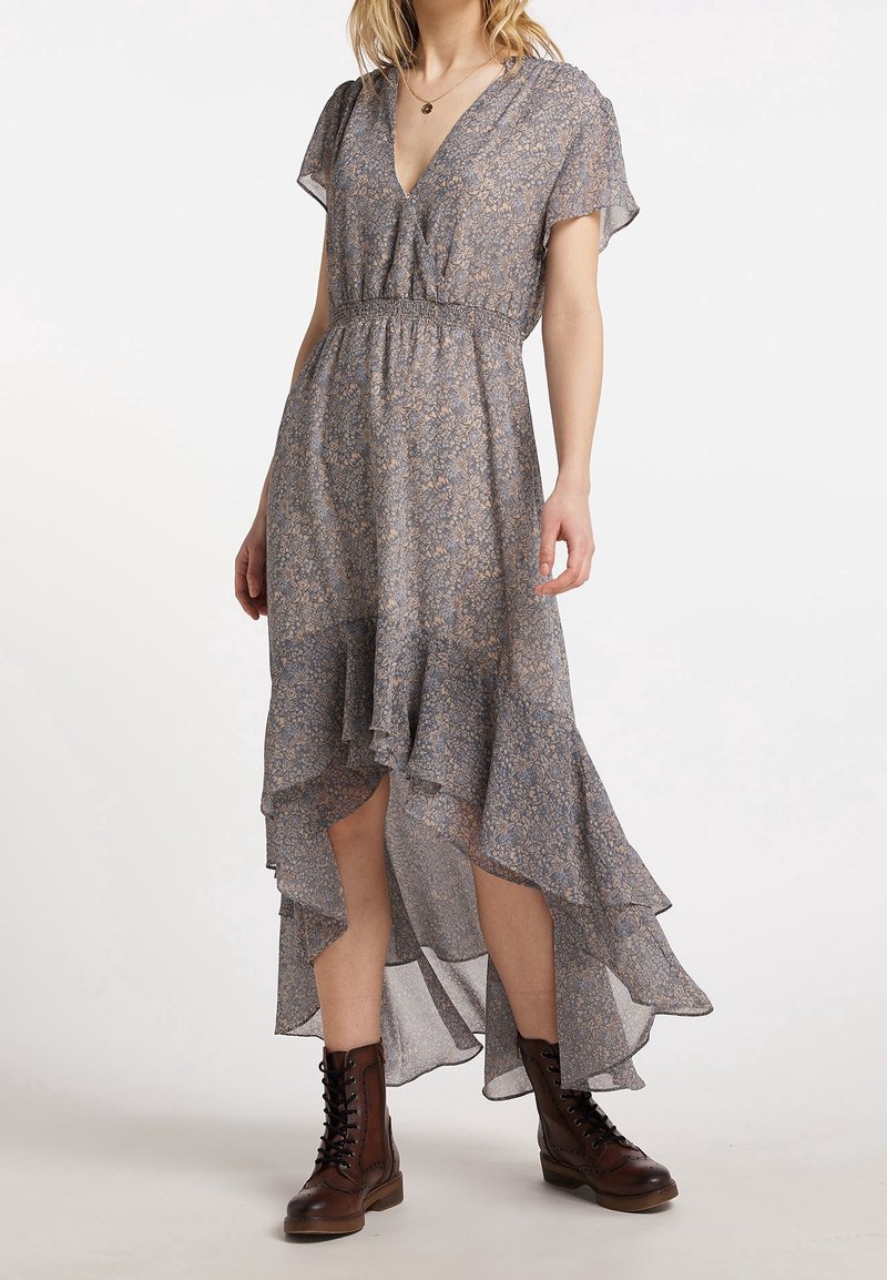 Femme portant une robe fluide à fleurs de style asymétrique, à manches courtes, encolure en V, taille smockée, et des bottines marron à lacets jusqu'aux chevilles.