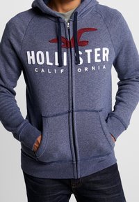 Huvad zip-up sweatshirt i marinblått texturerat tyg med vit "HOLLISTER CALIFORNIA" logotyp och röd acccent, som har framfickor och ribbade muddar.