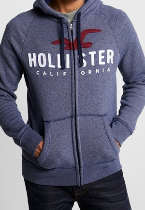 Kapuzensweatshirt mit Reißverschluss aus strukturiertem navyblauem Stoff mit weißem "HOLLISTER CALIFORNIA"-Logo und roten Akzenten, ausgestattet mit Fronttaschen und gerippten Bündchen.