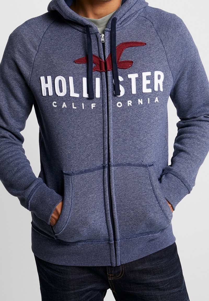 Huvad zip-up sweatshirt i marinblått texturerat tyg med vit "HOLLISTER CALIFORNIA" logotyp och röd acccent, som har framfickor och ribbade muddar.