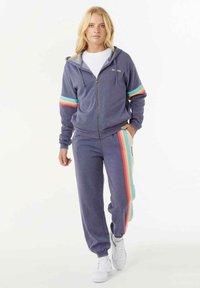 Paarse fleece zip-up jas met turkooise en koraalstrepen op de mouwen, gecombineerd met bijpassende sweatpants met zijstrepen. Witte sneakers.