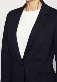 Mujer vistiendo un blazer negro con solapas de pico sobre una camiseta blanca de cuello redondo con un texto tenue en el pecho.