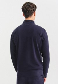 Granatowy sweter z półgolfem, gładką fakturą i ściągaczami na rękawach. Charakteryzuje się luźnym krojem i prostym wykończeniem na wysokości talii.