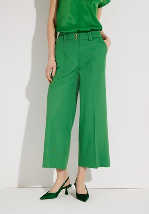 Femme portant un pantalon vert à taille haute et jambes larges avec une taille à double bouton et des escarpins verts à bride arrière, main dans la poche, sur fond uni.