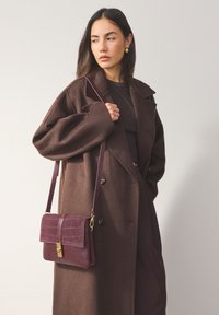 Sac à main bordeaux avec motif crocodile en relief, ferrures dorées et bandoulière, assorti à un manteau marron oversize.