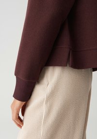 Persoon in een donkerbruine top met lange mouwen en geribbelde manchetten, met beige corduroy broek, staand met een ontspannen hand aan de zijde.