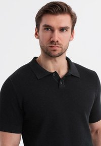 Polo negro de manga corta con un tejido texturizado, cuello plano y tapeta de dos botones. Diseño sencillo sin patrones ni acentos.