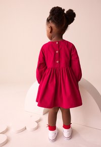 Vestito di cotone rosso con maniche lunghe a sbuffo, scollatura rotonda e chiusura con bottoni sul retro. Abbinato a sneakers bianche e calzini rossi.