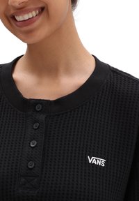 Zwarte trui met wafeltextuur, ronde hals, vijf zwarte knopen en een witte geborduurde "VANS" logo aan de linkerkant.