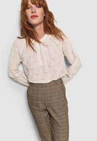 Vrouw met rood haar, gekleed in een witte blouse met eyelet en hoge, bruine geruite broek, staand met haar handen achter haar rug.