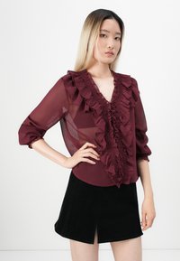 Burgunderrød bluse med volangdetaljer, gennemsigtig stof og dyb V-udskæring. Sammen med en sortvelour nederdel med slidser foran.
