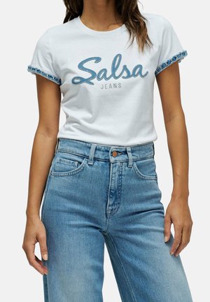 Vrouw draagt wit T-shirt met denimbies en de tekst "Salsa Jeans", gecombineerd met lichtblauwe high-waisted denim jeans.