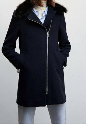 Femme portant un manteau zippé bleu marine avec un col en fourrure noire, un col roulé blanc, une chemise bleue et un jean bleu clair, les mains dans les poches du manteau.