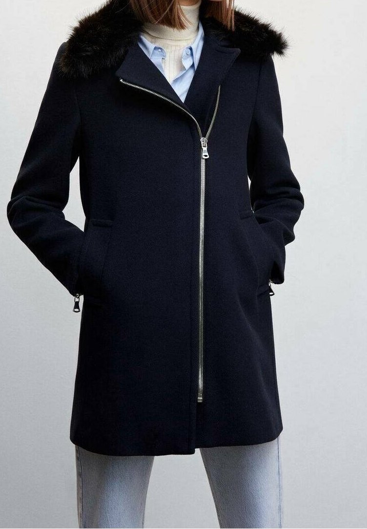 Femme portant un manteau zippé bleu marine avec un col en fourrure noire, un col roulé blanc, une chemise bleue et un jean bleu clair, les mains dans les poches du manteau.