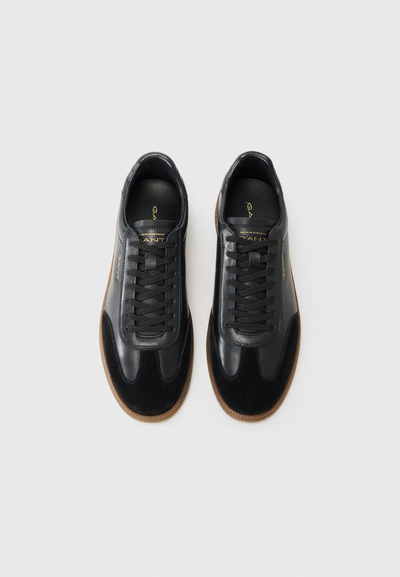 Baskets en cuir noir avec des accents en daim, bout rond, lacets plats et semelle en caoutchouc brun. Marque dorée visible à l'intérieur de la chaussure.