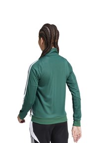 Giacca sportiva verde con colletto alto, maniche lunghe e strisce bianche sui lati. Il tessuto sembra liscio ed elastico, progettato per l'abbigliamento attivo.