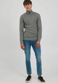 Suéter gris con cuello con cremallera y patrón en chevrón, puños y dobladillo acanalados. Combinado con jeans azules y zapatillas negras.