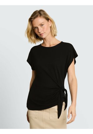 Vrouw met kort blond haar, die een zwart topje met korte mouwen draagt dat in de taille is gestrikt, en een beige rok tegen een lichte achtergrond.