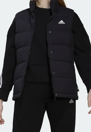Frau trägt eine schwarze Adidas-Steppweste über einem schwarzen Sweatshirt und Hosen mit Adidas-Logos, steht mit leicht ausgestreckten Armen.