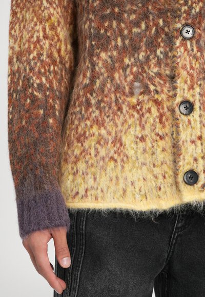 Kollane, oranž ja pruun niidist kardigan, millel on täpiline tekstuur, mustade nööpide ja lillaka rannekätiga. Kandmine tumedate teksade peal.