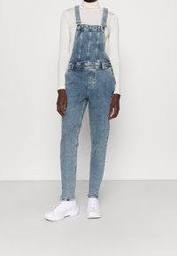 Vaaleansiniset farkkuhaalarit, joissa on säädettävät olkaimet, etutasku ja kapeneva lahje, puettuna valkoisen poolopaitaan.