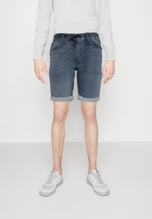 Personne portant un short en jean foncé retroussé avec une taille à cordon, une chemise blanche à manches longues et des baskets gris clair, debout sur un sol gris.